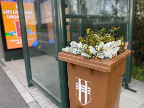 Brún tunna full af gróðri við strætóskýli