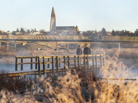 Tvær manneskjur á gangi í Vatnsmýrinni í vetrarstillum. Hallgrímskirkja og göngubrúin yfir Miklubraut í baksýn.