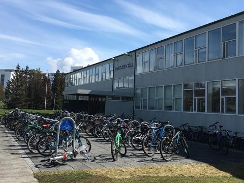Framhlið Hagaskóla, hjólastandar fyrir utan og mörg reiðhjól. Blár himinn, grænt gras, gott veður.