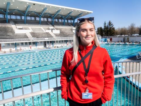 Dagbjört Ýr Gísladóttir, vaktstjóri í Laugardalslaug. Stendur á brúnni yfir laugina, í rauðri peysu með sólgleraugu á enni. Heiðskýrt úti og stúkan í baksýn.