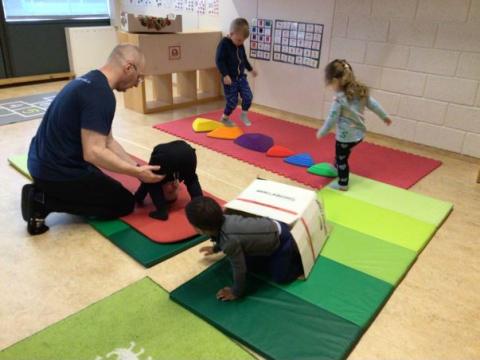 Börn á Bakkaborg í íþróttatíma. Eitt barn fær aðstoð við kollhnís, eitt skríður í gegnum pappagöng og tvö tipla á litríkum dýnum.