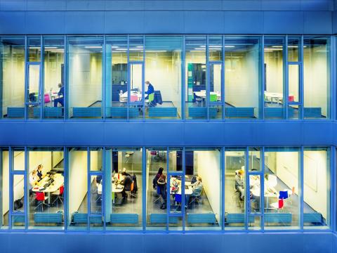 Séð inn um glugga þar sem fólk er að vinna við tölvur.