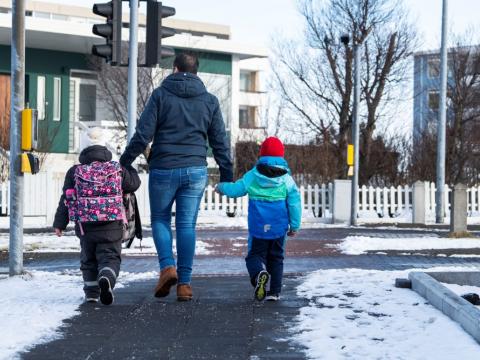 Maður og tvö börn á leið að ganga yfir götu, horft aftan á þau. Snjór úti, annað barnanna með skólatösku.