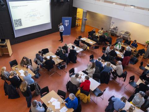 Fólk situr við borð í sal Vogaskóla og borgarstjóri stendur við skjá