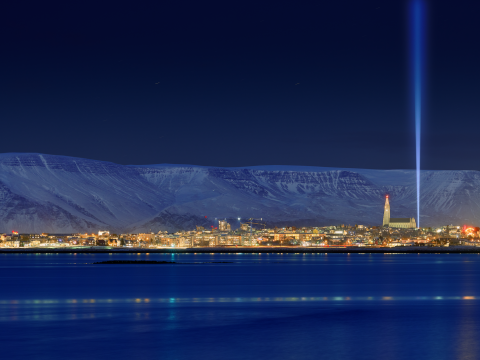 Imagine Peace Tower. Night view of the city with Mount Esja in the background and water in the foreground. Blue tones, with a column on the far right.