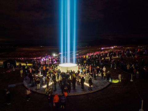 Friðarsúlan tendruð í Viðey. Myrkur, fullt af fólki í kringum súluna, sem lýsir upp í svartan himinn.