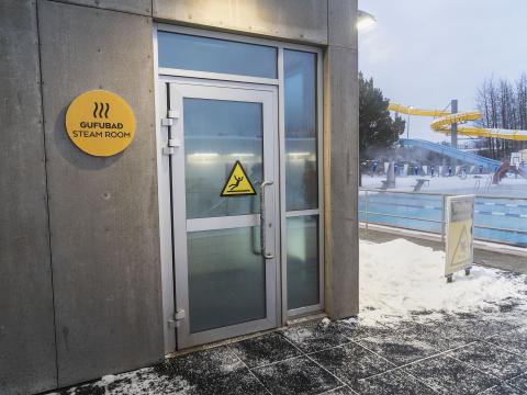 Sauna at Breiðholtslaug, showing the exterior, entrance door, and sign next to the door. Swimming pool in the background. Winter, snow.