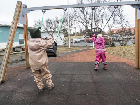 Tvö leikskólabörn á leikskólalóð að leika sér í rólum. Eru í pollagalla, halda í sitthvora róluna og snúa baki í myndavélina.