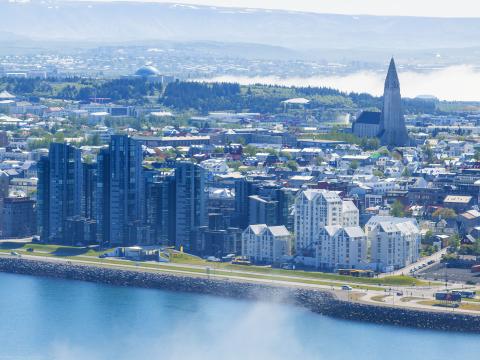 Aerial photo of Reykjavík, view from Sæbraut across the city center toward Hallgrímskirkja church and Perlan. Ocean in the foreground, mountains and sea in the background. Mist overhead.