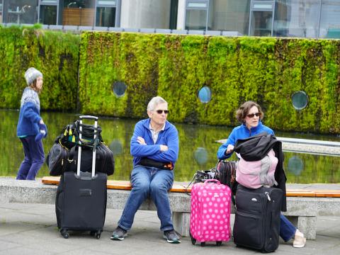 Tourists, a man and woman, sit with travel bags by the pond at City Hall. Young woman with the moss wall of City Hall in the background.