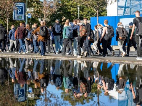 A group of youth, seen from behind, walking by water, their images reflected on the surface.