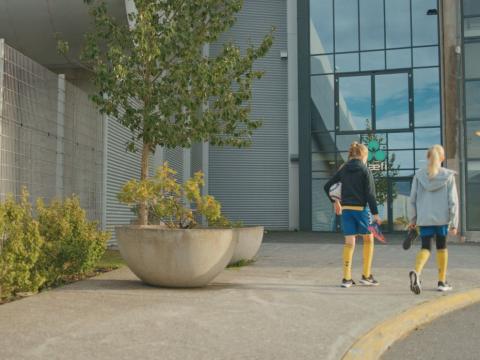 Tvær ungar stúlkur í fótboltaæfingafötum labba frá myndavélinni að byggingu. Þær eru hægra megin í mynd, vinstra megin eru gróðurker. Sumar.