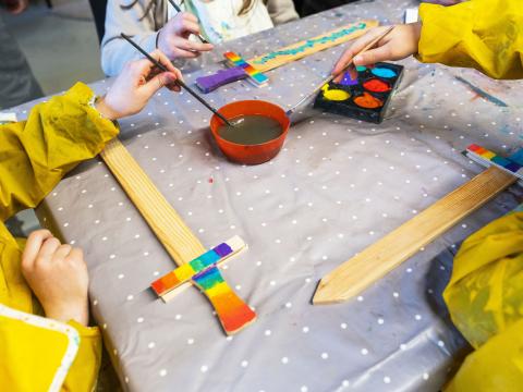 Children sit at a table crafting. We can only see their hands in yellow protective clothing. A sword being painted.
