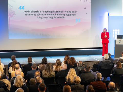 Mayor Heiða Björg Hilmisdóttir in a red suit. She stands at the far right of the stage while addressing a large audience in Tjarnarsalur. Housing meeting. Text on a banner behind her.