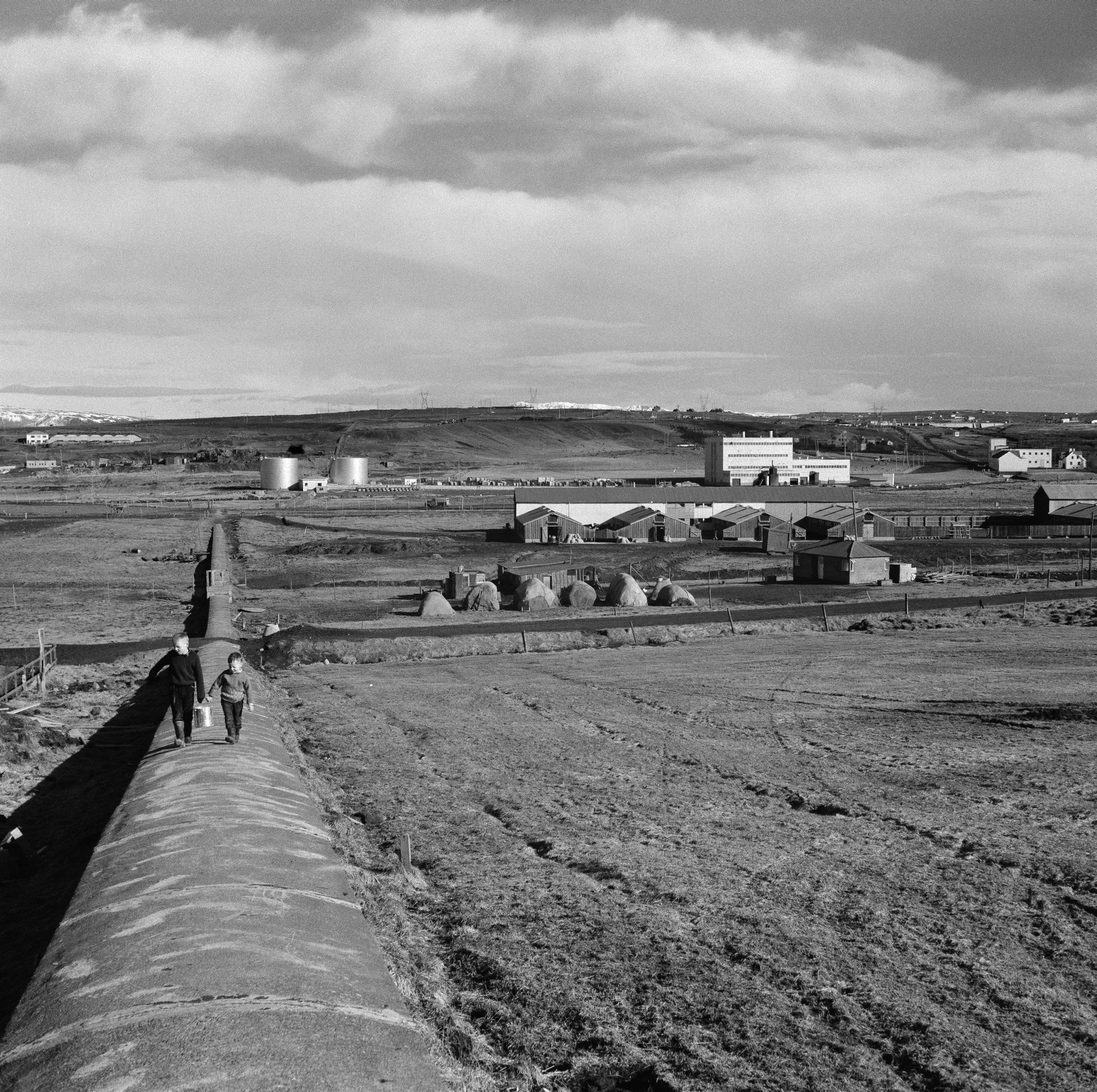 Árið 1963: Skeiðvöllur, hesthús og hlaða hestamannafélagsins Fáks við Elliðaár. Fjær er Toppstöðin, gamla rafstöðin og fleiri byggingar í Elliðaárdal. Tveir drengir koma gangandi upp steinsteyptan hitaveitustokk með fötu á milli sín. Í fjarska til vinstri sjást kartöflugeymslurnar í Ártúnsbrekku. Mynd frá Ljósmyndasafni Reykjavíkur.