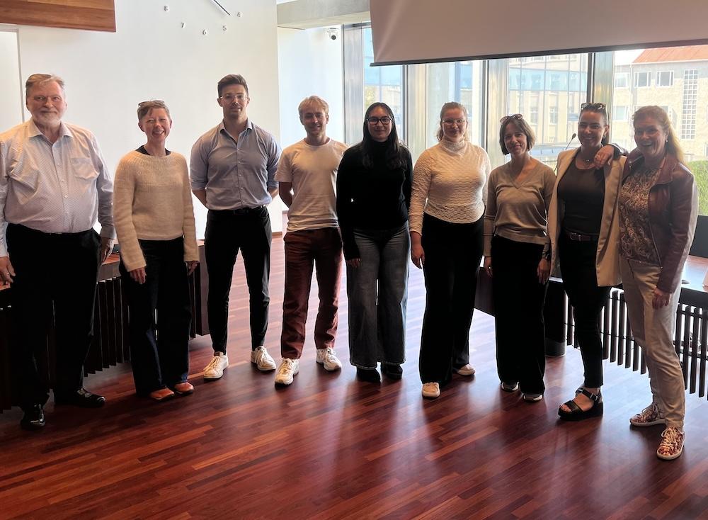 People in a row inside a hall with parquet floors and windows.