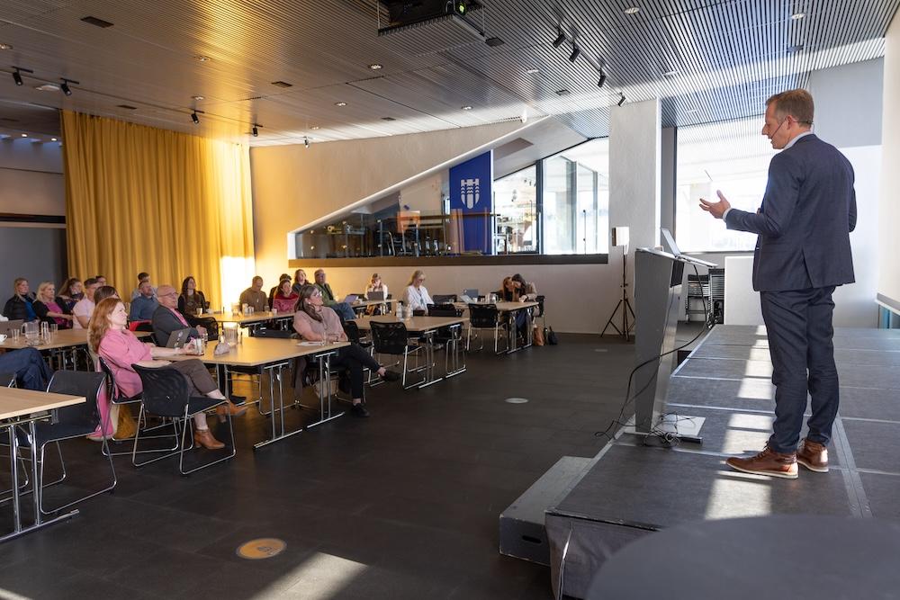 Man speaking on stage and looking over people at tables in the hall.