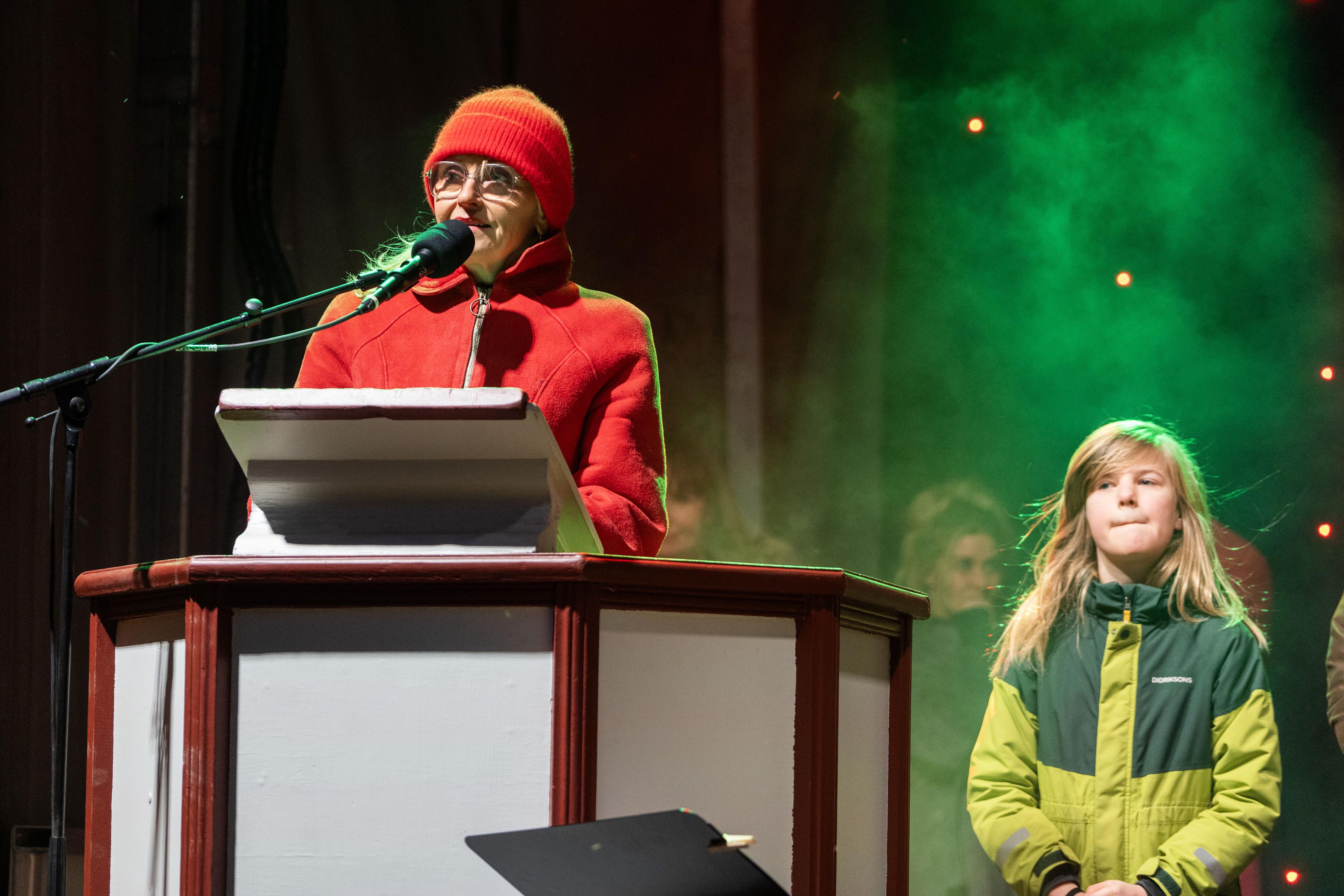 A woman in red speaks at the podium, with a young boy standing beside her.
