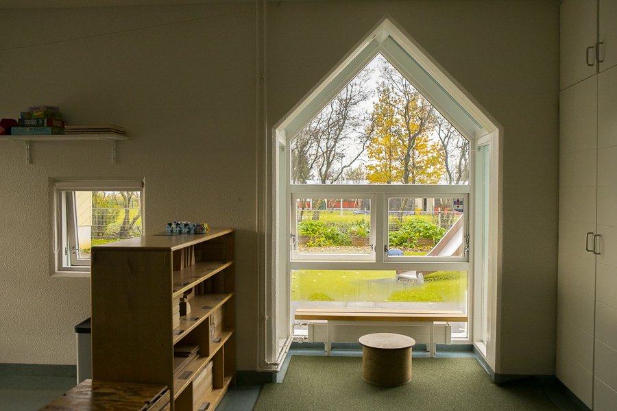 Séð út um glugga á leikskólanum Sólborg. Inni er hilla og lítill stóll en úti rennibraut og önnur leiktæki.