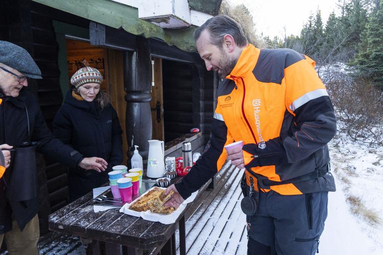 Einar Þorsteinsson borgarstjóri þiggur veitingar við skála félags Norðmanna á Íslandi í Heiðmörk. Snjór, bakkelsi á borði á palli hússins.