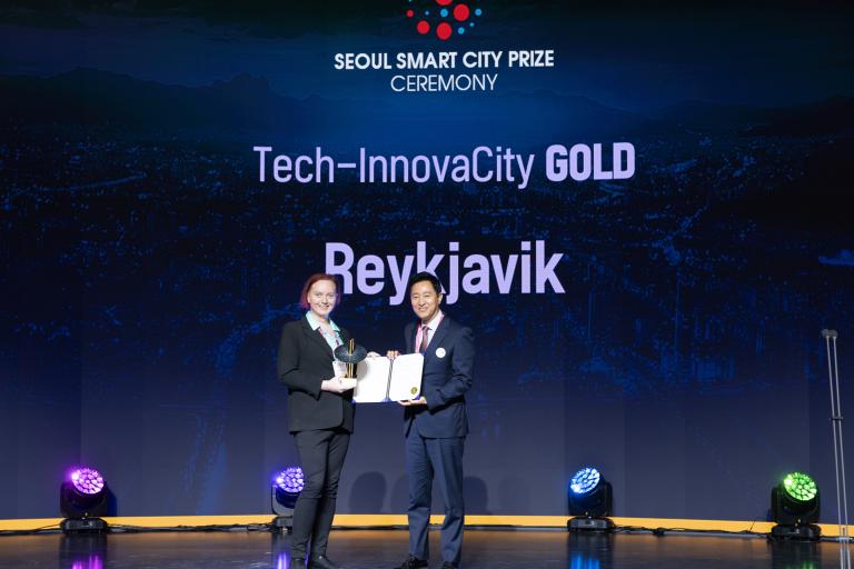 Alexandra Briem á sviði í Seoul að veita viðtöku Smart City Awards. Maður afhendir henni verðlaun og á bakvið þau stendur Tech-InnovaCity GOLD Reykjavík