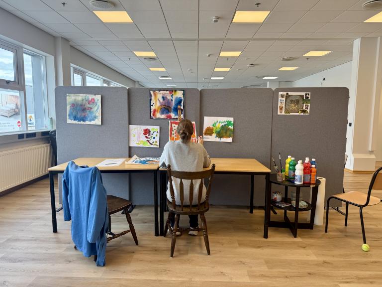 A woman sits at a table in front of a partition decorated with pictures. 