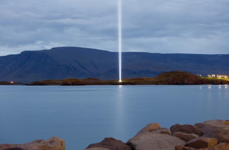 The Imagine Peace Tower in Viðey, likely viewed from Skarfabakki or Sæbraut. The tower is seen at dusk, with Mt. Esja in the background.