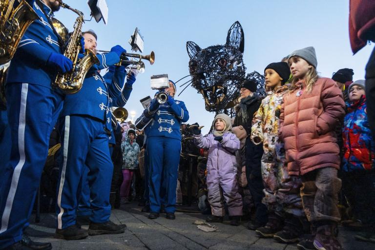 From The Yule Cat lighting ceremony at Lækjartorg. Photo taken from ground level shows brass band members playing on the left, the cat's head on the right and children watching the band.