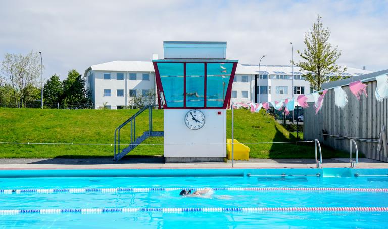 Lifeguard tower at Vesturbæjarlaug. Pool in the foreground with a person swimming. Green grass, summer