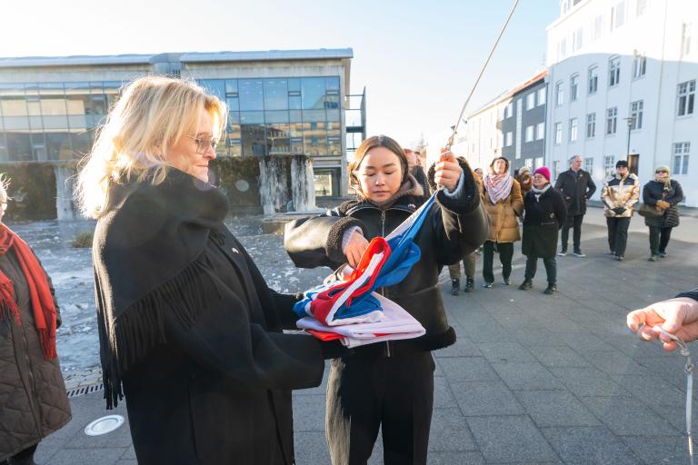 Heiða Björg Hilmisdóttir borgarstjóri og Malu Hendriksen, grænlenskur starfsnemi á sendiskrifstofu Grænlands á Íslandi. Þæ halda á íslenska fánanum, reiðbúnar að draga hann að húni. Skömmu síðar var sá grænlenski dreginn að húni. Fólk í bakgrunni, blár himinn, vetur.