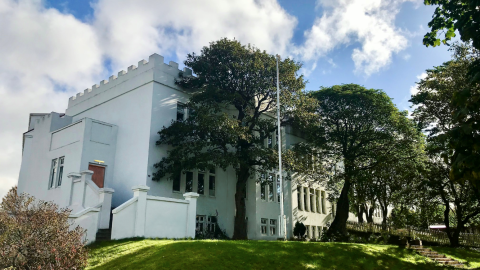 Hús Söngskólans í Reykjavík séð utanfrá. Stór tré í garðinum, hvítt hús og blár himinn.