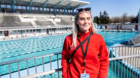 Dagbjört Ýr Gísladóttir, vaktstjóri í Laugardalslaug. Stendur á brúnni yfir laugina, í rauðri peysu með sólgleraugu á enni. Heiðskýrt úti og stúkan í baksýn.