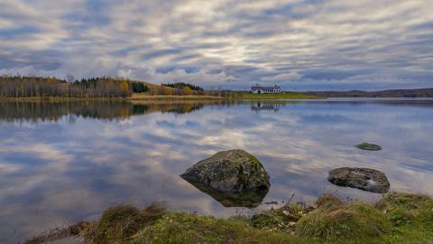 Elliðavatn í logni, skýin speglast í vatninu og í forgrunni eru stórir steinar í vatninu. Elliðabærinn í bakgrunni hinum megin við vatnið.