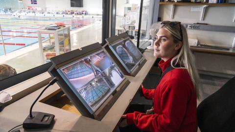 Lifeguard Dagbjört Ýr Gísladóttir monitors screens in the guard room at Laugardalslaug Pool. She's wearing a red sweater, with a window overlooking the indoor pool on the left side of the image.