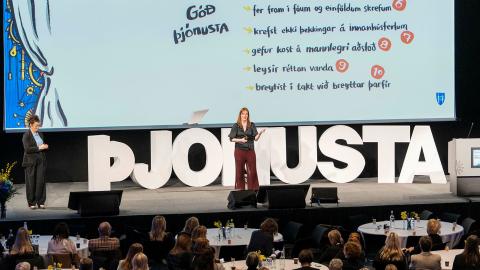 Arna Ýr Sævarsdóttir, Head of Office for services and transformation at the Department of Service & Innovation, at Reykjavík City's management day at Harpa 2024. Arna center stage, with the letters ÞJÓNUSTA prominent on the stage and slides in the background. Seated guests in the foreground.