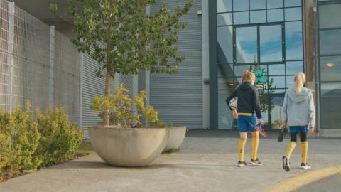 Tvær ungar stúlkur í fótboltaæfingafötum labba frá myndavélinni að byggingu. Þær eru hægra megin í mynd, vinstra megin eru gróðurker. Sumar.