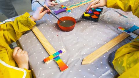 Children sit at a table crafting. We can only see their hands in yellow protective clothing. A sword being painted.