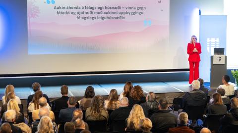 Mayor Heiða Björg Hilmisdóttir in a red suit. She stands at the far right of the stage while addressing a large audience in Tjarnarsalur. Housing meeting. Text on a banner behind her.